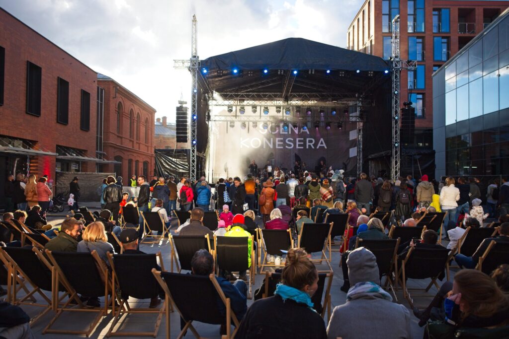 Koncert na dziedzińcu Centrum Praskiego Koneser, ludzie siedzą na leżakach, inni stoją przed sceną, na której gra zespół, dzień, jesień.