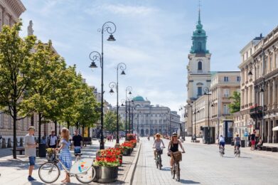 Krakowskie Przedmieście, ludzie jeżdżą na rowerach, inni spacerują, w tle Pałac Staszica, Bazylika Świętego Krzyża, po lewej stronie wzdłuż ulicy rosną drzewa, słoneczny dzień, lato.