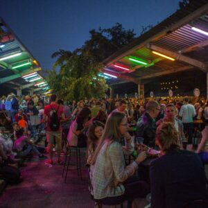 Nocny Market na Dworcu Głównym, ludzie siedzą przy stolikach, jedzą, piją, rozmawiają, w tle stoiska z jedzeniem, noc, lato.