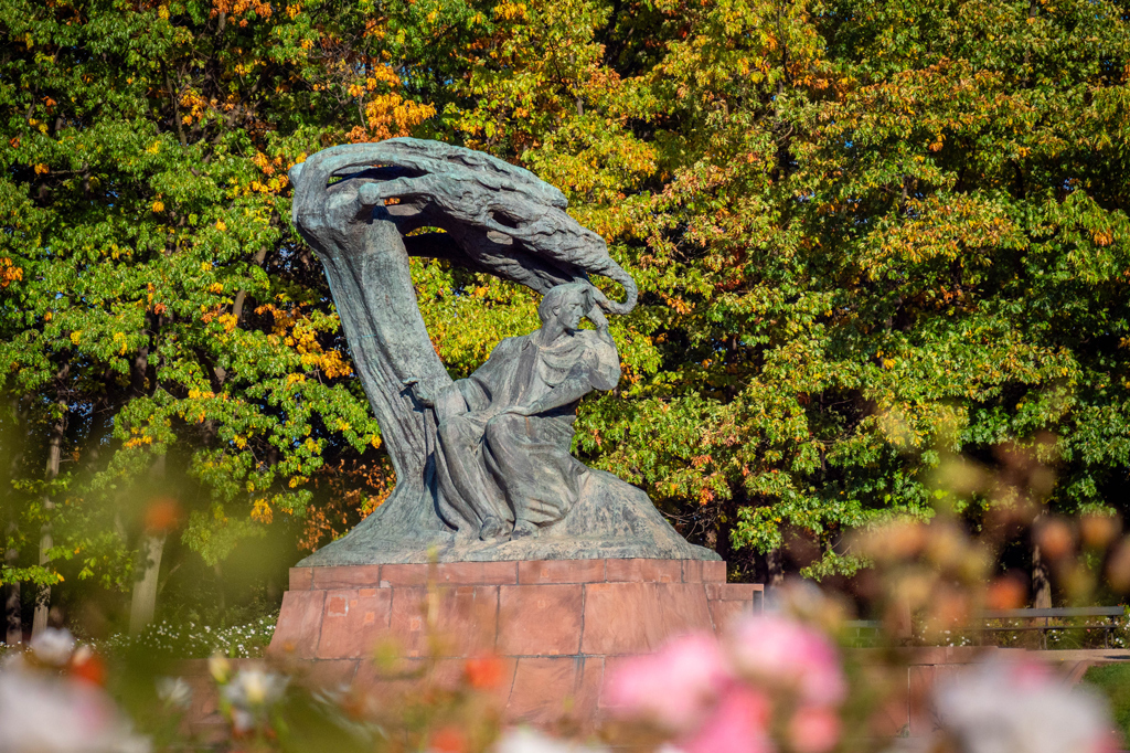 Pomnik przedstawiający mężczyznę w powłóczystych szatach siedzącego pod konarem drzewa z pochylonymi gałęziami. Za pomnikiem drzewa pokryte liśćmi w zielonych i pomarańczowych kolorach.